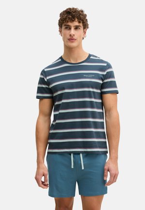 Man met krullend haar, gekleed in een marineblauw, wit en rood gestreept T-shirt en groene shorts met een trekkoord, tegen een witte achtergrond.