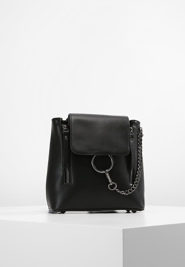 Sac à main en cuir noir avec rabat avant, détail anneau métallique, deux fermetures éclair verticales et une bandoulière chaîne posé sur une surface blanche.