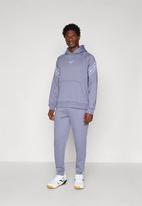 Γκρι αθλητική μπλούζα με κουκούλα και μπροστινή τσέπη, ταιριαστό παντελόνι φόρμας και λευκά παπούτσια με κίτρινες λεπτομέρειες. Διακοσμημένο με λογότυπα στα μανίκια.