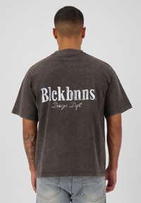 Donkerbruine oversized t-shirt met korte mouwen, met op de achterkant in witte tekst "Blckbnns Design Dept.". Vervagende, iets gestructureerde stof.