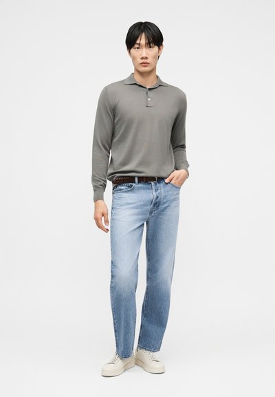 Νέος άνδρας στέκεται φορώντας γκρι πόλο με μακριά μανίκια, ανοιχτόχρωμο μπλε τζιν με καφέ ζώνη και λευκά αθλητικά παπούτσια, με τα χέρια χαλαρά στα πλάγια.