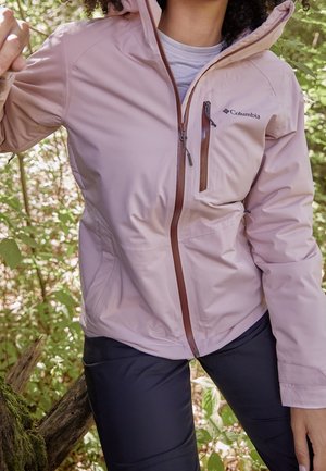 Osoba ubrana w jasnoróżową kurtkę Columbia i ciemne spodnie, stojąca na zewnątrz wśród zielonych liści, trzymająca gałąź jedną ręką.