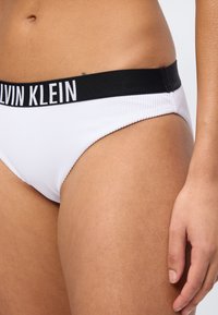 Krupni plan osobe koja nosi bijeli Calvin Klein donji rublje s crnim pojasom i logotipom, prikazujući torzo, bok i ruku.