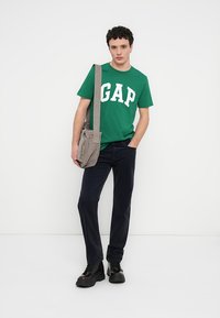 Πράσινο βαμβακερό t-shirt με λευκό λογότυπο "GAP", συνδυασμένο με σκούρο μπλε παντελόνι και γκρι τσάντα ώμου με υφή. Μαύρα μποτάκια ολοκληρώνουν την εμφάνιση.
