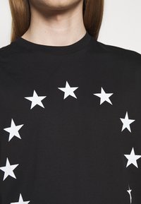 Černé bavlněné tričko s kruhovým vzorem bílých hvězd. Kulatý výstřih, krátké rukávy a hladká, rovnoměrná textura.