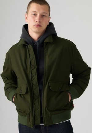 Jeune homme portant un sweat à capuche noir sous une veste vert foncé ouverte, les mains dans les poches, fond neutre.