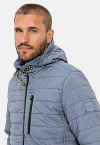 Lyseblå quilted jakke med hætte, sort lynlås og guld snoredetaljer. Indeholder en lynlåslomme på brystet og tekstureret stof.