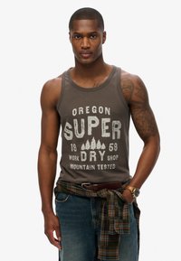 Bruine tanktop met witte bedrukte tekst en een boomgrafiek. Gemaakt met blauwe spijkerbroek en een geruite shirt om de taille gebonden. Donkere huid, kort haar.