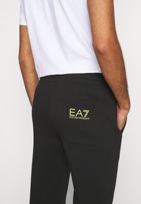 Černé joggingové kalhoty s měkkou texturou, vybavené zadní kapsou a neonově zeleným logem EA7 Emporio Armani. Jednoduchý, ležérní design.