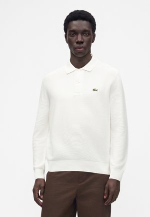 Uomo con capelli corti e ricci che indossa una polo bianca a maniche lunghe con logo verde e pantaloni marroni su uno sfondo chiaro e uniforme.
