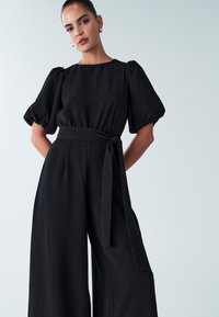 Zwarte jumpsuit met pofmouwen en een aansluitend lijfje. Voorzien van wijde broekspijpen en een stoffen riem gestrikt om de taille. Gladde textuur.