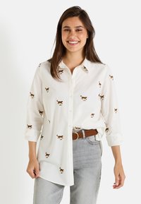 Chemise blanche à boutons en coton avec des motifs de chats marron brodés. Présente un col et des manches retroussées. Associée à un jean gris.