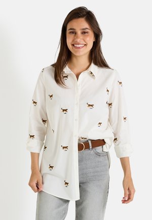 Chemise blanche à boutons en coton avec des motifs de chats marron brodés. Présente un col et des manches retroussées. Associée à un jean gris.