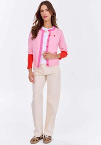 Różowy i czerwony kardigan z zapięciem na guziki, ze szczegółem w kształcie serca, kontrastującymi rękawami, noszony na białej bluzce, zestawiony z jasnymi spodniami i wzorzystymi butami.