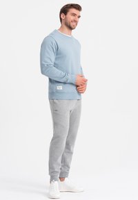 Pullover blu chiaro in tessuto morbido con polsini e orlo a coste, abbinato a pantaloni della tuta grigi e sneakers bianche, dotato di tasche laterali.