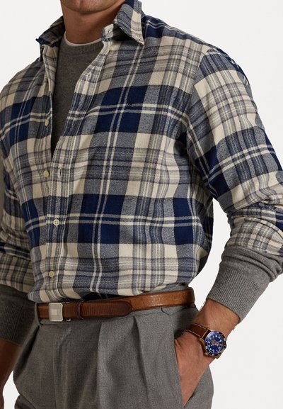 Blauw en beige geruite shirt met knoopsluiting aan de voorkant, grijze trui eronder, grijze broek, bruine leren riem en een blauwe horloge om de pols.