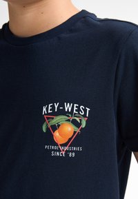 Tricou din bumbac de culoare albastru marin, cu un design grafic cu portocale și frunze, încadrat într-un triunghi roșu, și textul "KEY-WEST PETROL INDUSTRIES DIN '89."