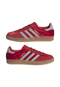 Červené semišové sportovní boty s třemi úzkými bílými pruhy, zlatým nápisem "Gazelle" a průhlednou gumovou podrážkou.