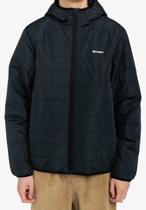 Personne portant une veste matelassée noire zippée à capuche avec un petit logo blanc Element sur la poitrine et un pantalon beige.