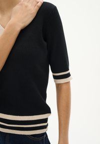 Schwarzer gerippter Pullover mit kurzen Ärmeln, der beige gestreifte Akzente an den Ärmeln und am Saum aufweist. Der V-Ausschnitt verleiht ihm eine moderne Note.