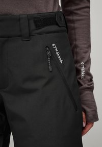 Black outdoor pants with a textured finish, featuring a front zip pocket, Velcro waistband, and white logo detailing on the side.