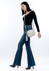 Kvinde går iført sort langærmet top, blå vidde jeans, sorte støvler med hæl og en hvid crossbodytaske med frynse-detalje.