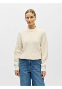 Kermanvärinen ribbineulottu neulepaita, jossa pitkät hihat, korkea kaulus ja rento istuvuus. Yhdistetty vaaleansinisiin leveälahkeisiin farkkuihin.