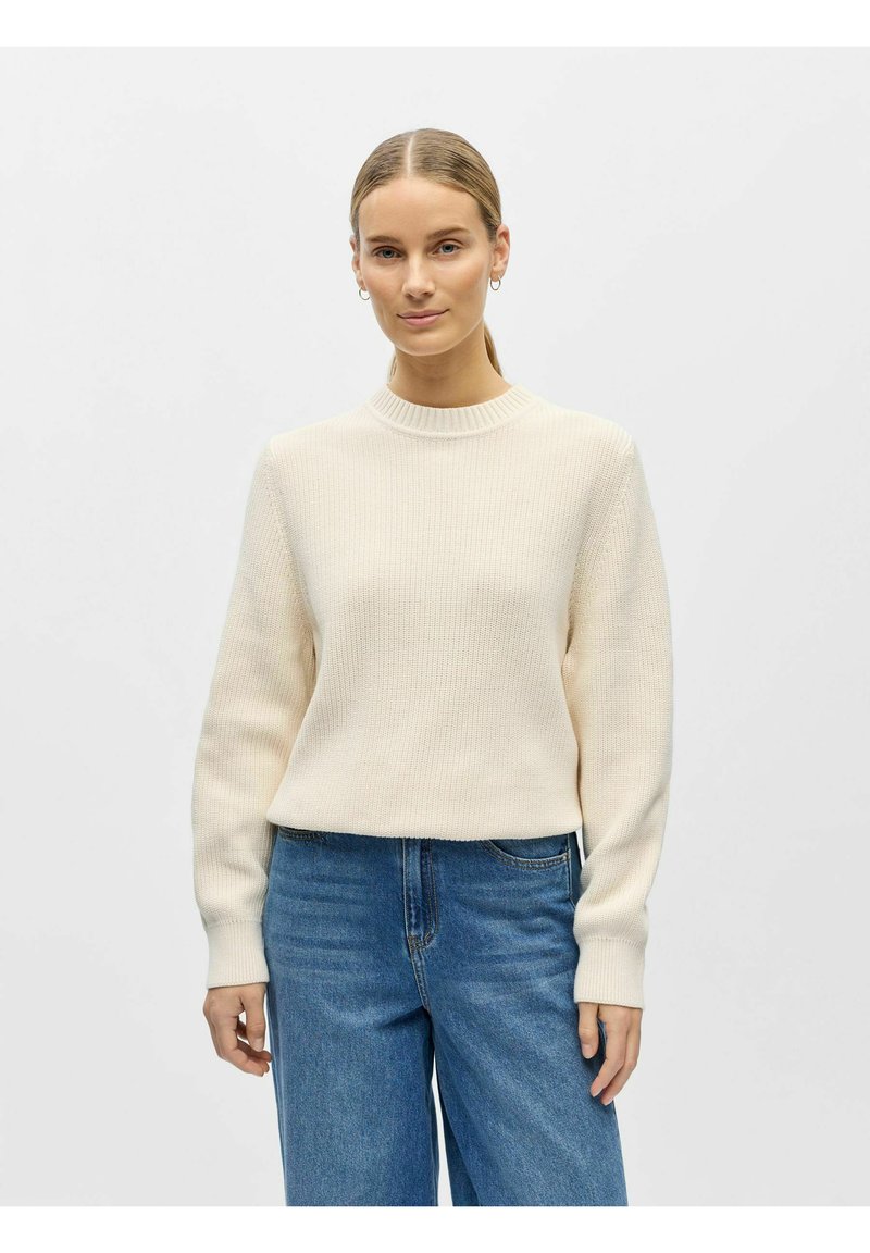 Kermanvärinen ribbineulottu neulepaita, jossa pitkät hihat, korkea kaulus ja rento istuvuus. Yhdistetty vaaleansinisiin leveälahkeisiin farkkuihin.