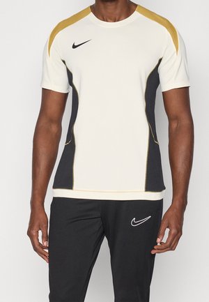 Man draagt een wit en goud Nike sportshirt met korte mouwen, zwarte zijpanelen, en zwarte Nike broek.