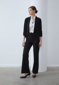 Schwarze, maßgeschneiderte Hose kombiniert mit einer weißen Bluse mit Schleife, die unter einem schwarzen Cardigan mit offenem Front getragen wird. Glatte Texturen und elegantes Design.