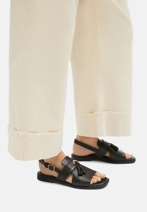 Sandalias negras de cuero con suela plana, que presentan bandas anchas y detalles de borlas. Combinadas con pantalones crema enrollados.