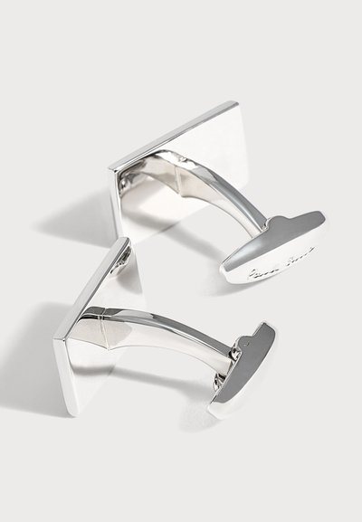 Paire de boutons de manchette rectangulaires en argent brillant avec des barres de liaison courbées, posée sur une surface blanche avec des ombres douces.