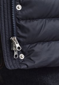 Marineblå dunjakke med en glat, skinnende tekstur. Har en sølvfarvet lynlås og trykknapdetalje, der fremhæver et quiltet design.