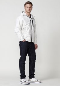 Lichte, witte, ritssluitingjas met een capuchon en zwarte accenten. Gecombineerd met donkerblauwe jeans en witte sneakers met grijze details.