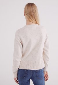 Lichtbeige sweater van gebreide textuur met gespikkelde accenten. Heeft een ronde halslijn en ribgebreide manchetten en zoom. Draagt met blauwe jeans.