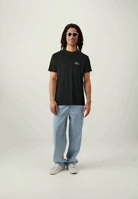 Black cotton t-shirt with short sleeves and a small gray logo on the chest, paired with light blue jeans and white sneakers.