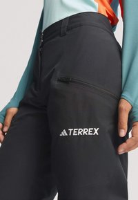 Čierne vonkajšie nohavice so hladkou textúrou, s bočnou zipsovou kapsou a vyšitým logom "TERREX". Nosia sa s modrým tričkom s dlhým rukávom.