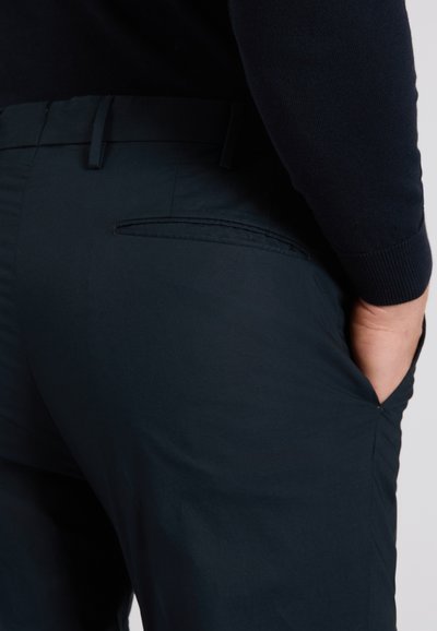 Uomo che indossa pantaloni blu scuro e una camicia a maniche lunghe nera, con la mano destra in tasca, visto di profilo dalla vita fino a metà coscia.