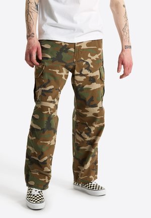 Uomo con pantaloni cargo mimetici marroni e verdi, maglietta bianca e scarpe slip-on a quadri nero e bianco. Tatuaggi visibili sugli avambracci.