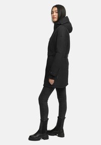 Parka nera con cappuccio, vita con cintura, tasche laterali con zip e tessuto texturizzato. Abbinata a jeans neri aderenti e stivaletti alla caviglia.