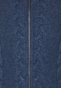 Närbild av marinblått stickat tyg med kabelmönster och en silverfärgad metallblixtlås som går ner längs mitten.