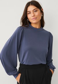 Marineblauw langslavige top met hoge hals, met pofschouders en een gladde textuur, gecombineerd met zwarte broeken.