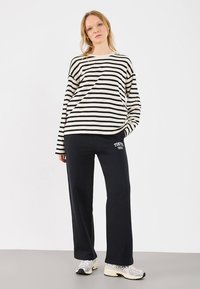 Camiseta de manga larga a rayas blancas y negras combinada con pantalones deportivos negros con el estampado "TOKYO 1953", usada con zapatillas deportivas grises.