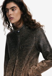 Veste en cuir synthétique marron avec un col haut, une finition texturée et une poche zippée sur la poitrine gauche. Présente une silhouette slim fit.