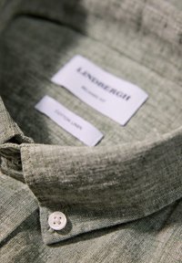 Zielona lniana koszula z kołnierzykiem, z białym guzikiem, z metkami marki "Lindbergh" i "Cotton Linen." Zbliżenie na teksturowany materiał.
