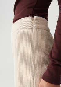 Close-up van beige corduroy broek met een zijrits, gedragen met een maroon langemouwtop, waarbij de textuur van de stof en de stikseldetails zichtbaar zijn.
