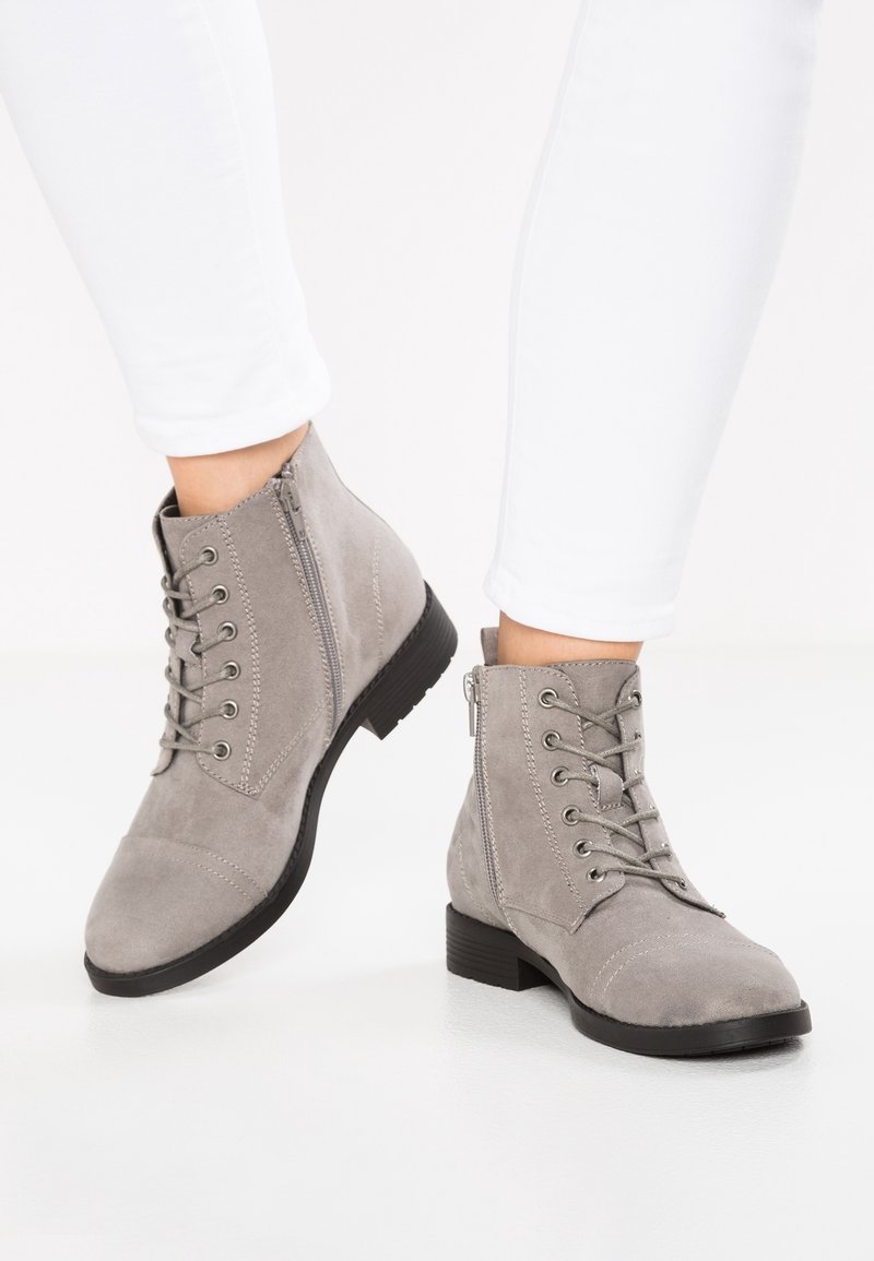 Stivali alla caviglia in suede grigio con lacci e zip laterali indossati con pantaloni aderenti bianchi su uno sfondo bianco.