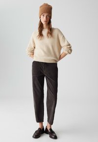 Cream knitted sweater, dark corduroy pants with pockets, and brown loafers. Model wears a tan beanie, standing with hands on hips.