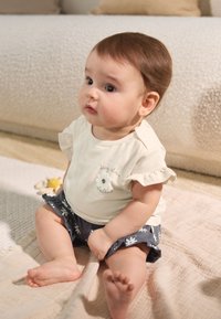 Hellbeiges Kurzarmhemd mit Rüschen und Blumenstickerei; marineblaue Shorts mit weißem Blumenmuster; sitzt auf einer strukturierten, neutralen Decke.