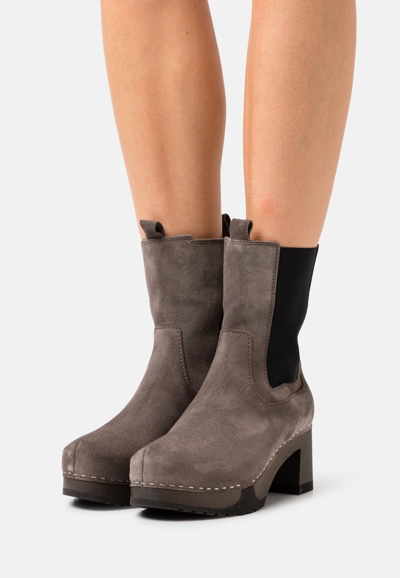 Jambes portant des bottines en daim gris à talons blocs avec des panneaux élastiques noirs sur les côtés, sur fond blanc.
